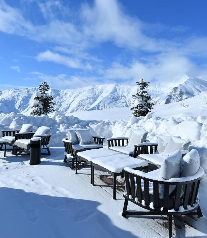 Snow-covered terrace with dark framed lounge chairs and low tables, snow-laden evergreens and a panorama of snow-capped ridges under a bright blue sky